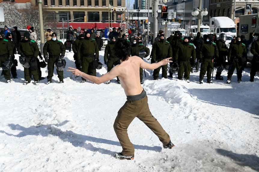 a shirtless man in brown trousers stands in the snow in front of a line of police. He is making the peace sign with both hands. 