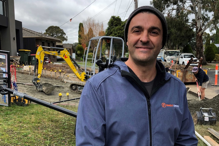 Matt Piccioli wearing a blue jacket and beanie, standing in front of a construction site with digger.