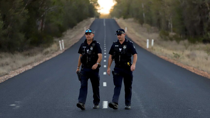Two police officers in Queensland uniform stand on a straight country road