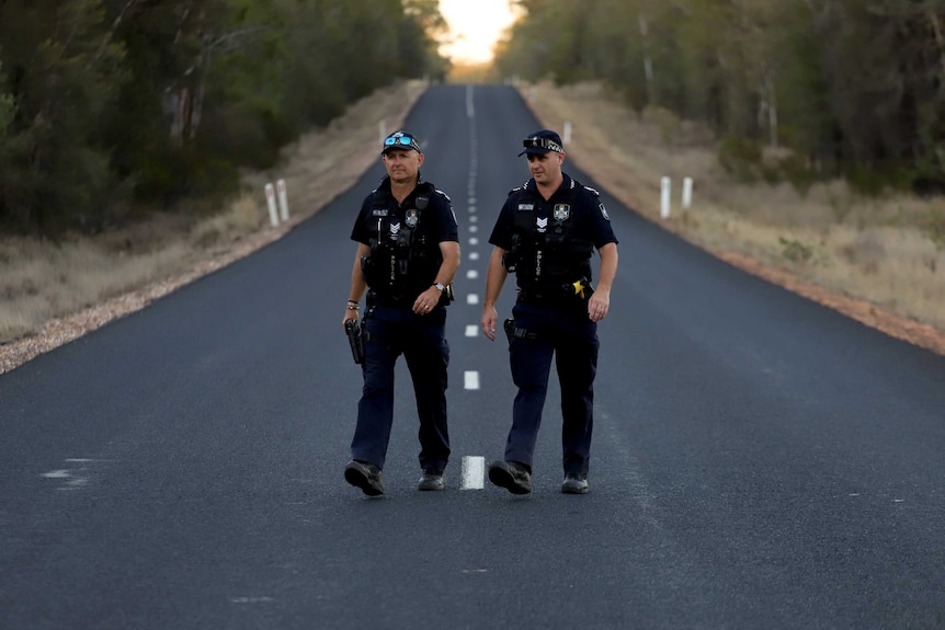 Two police officers in Queensland uniform stand on a straight country road