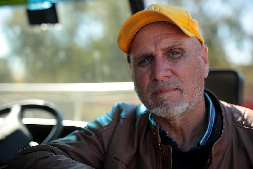 A man with grey stubble and wearing a yellow baseball cap leans against a bus.