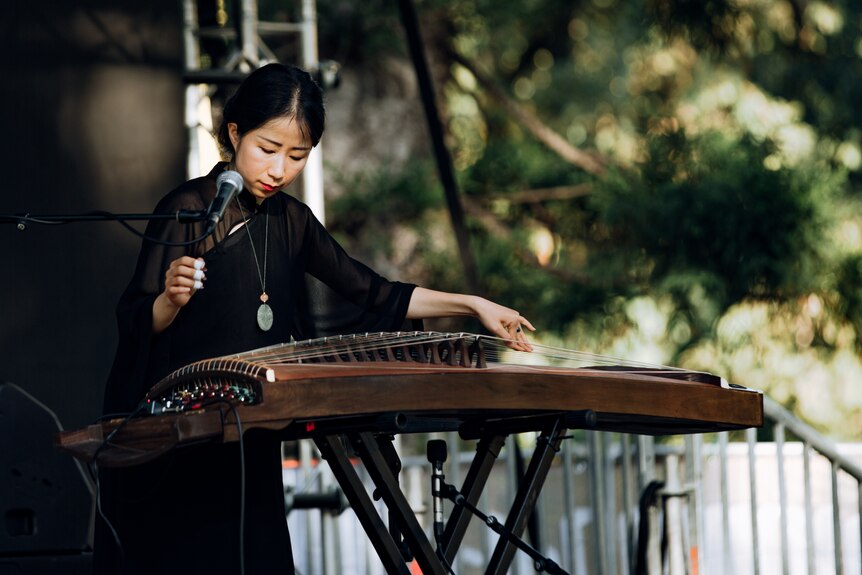 A woman in a black dress plays a guzheng on stage