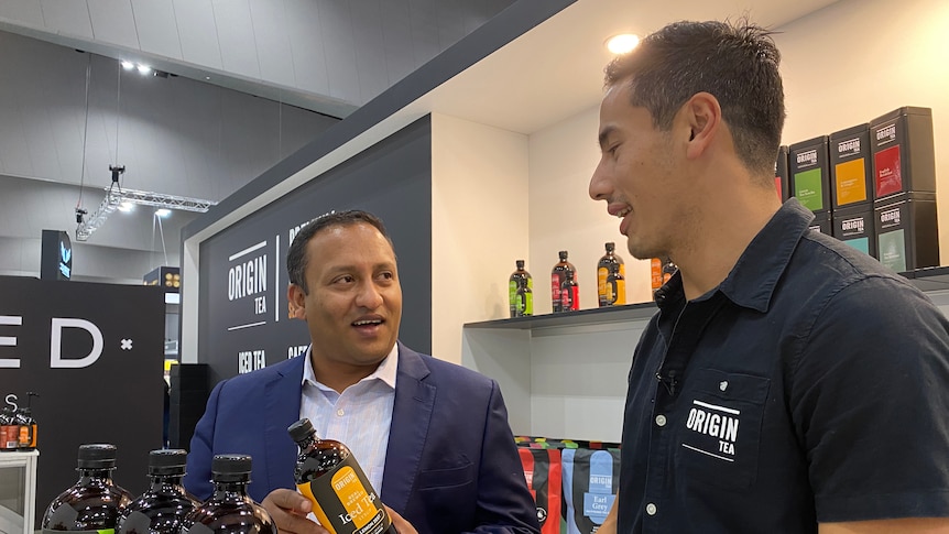 Two men, one in a black shirt and one in a blue suit are talking to each other about tea.