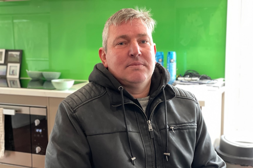 A man wearing a leather jacket and hoodie sits in front of a green wall in a kitchen.