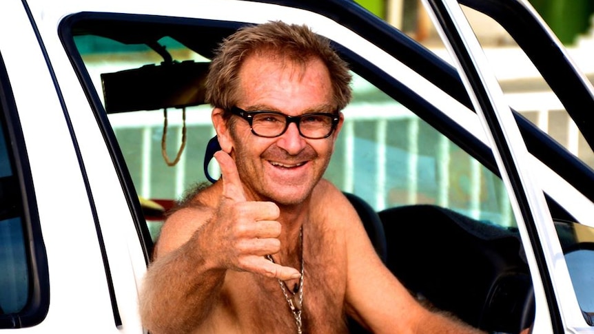 A man sits shirtless in a car doing a shakka hand signal
