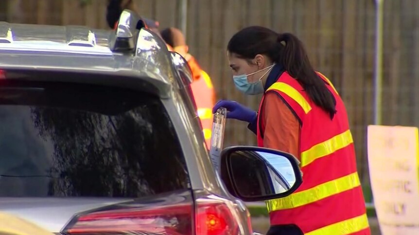 A woman in a high-vis vest and surgical mask holds up a clipboard as she speaks to the driver inside a silver car.