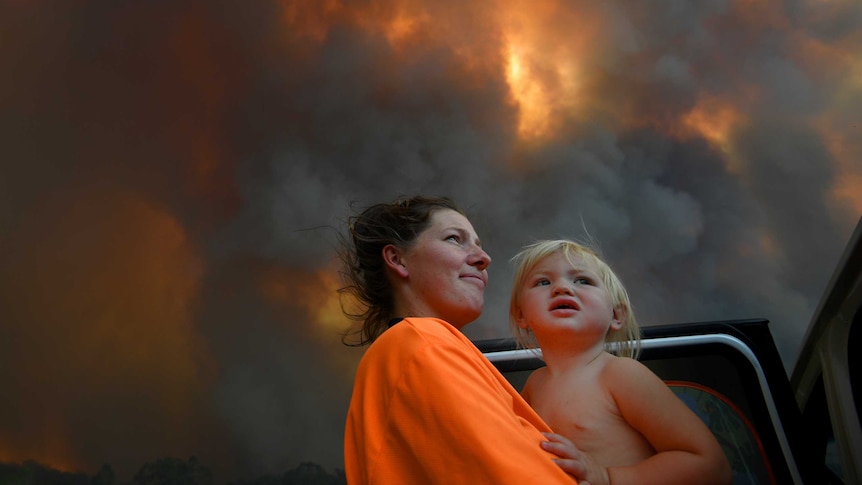 A woman and a young toddler look out a smoky, glowing sky while standing at an open car door.