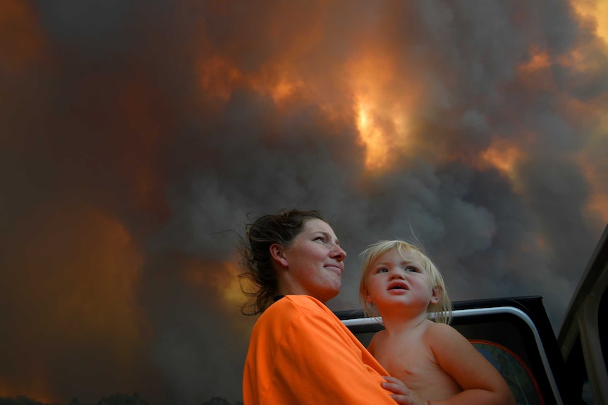 A woman and a young toddler look out a smoky, glowing sky while standing at an open car door.
