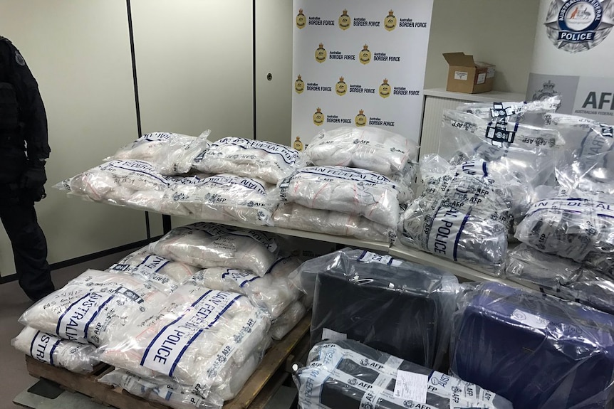 Large bags of methamphetamine crystals wrapped in police plastic are stacked up on each other and three suitcases sit in front.
