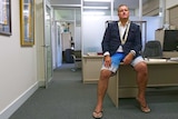 A man in a blazer and boardshorts with a mayoral chain around his neck sits looking moody in an office.
