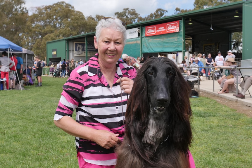 A woman kneeling down in a grassy area looking straight ahead with her pet Afghan hound 