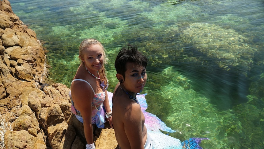 A woman and a man, both dressed as merfolk, sit on a rock at a swimming area, smiling.