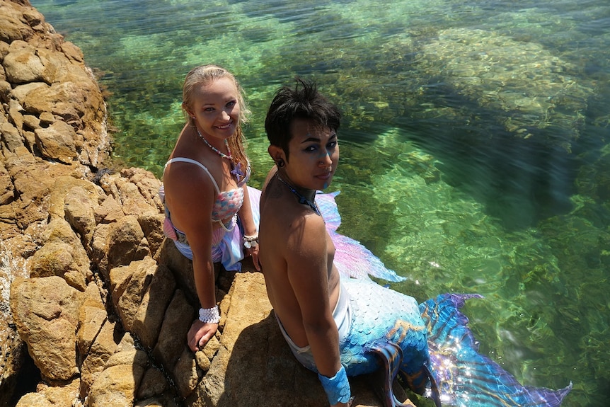 A woman and a man, both dressed as merfolk, sit on a rock at a swimming area, smiling.