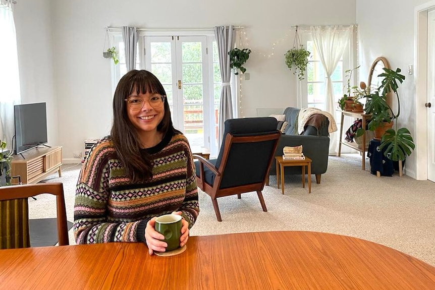 Jess Clarke sitting at a table in her Armidale home, drinking a cup of tea.