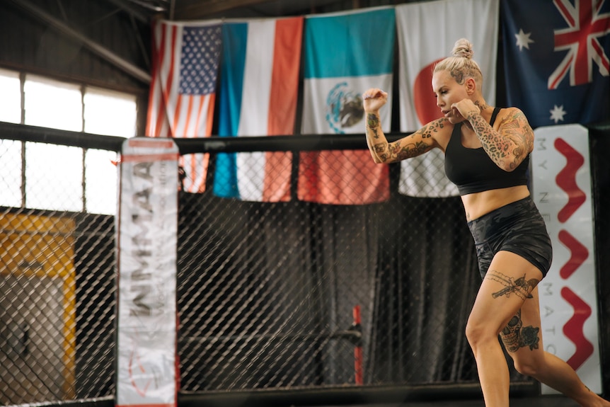  A woman with a tight ponytail wears boxing gloves in a boxing rink and is photographed mid punch.