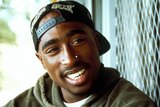 A portrait photo of Tupac Shakur. He is a black man in his 20s wearing a backwards cap. He is smiling.