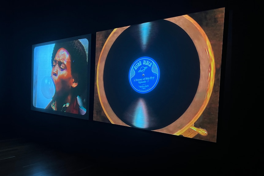 Two screens hang in a dark gallery space. On one screen is a young Black man blowing a bubble, on the other is a vinyl record. 