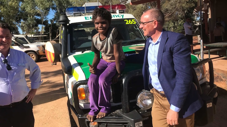 An Aboriginal boy sits on the hood of a 4-wheel-drive ambulance giving the camera the thumbs up. A man with glasses watches him.