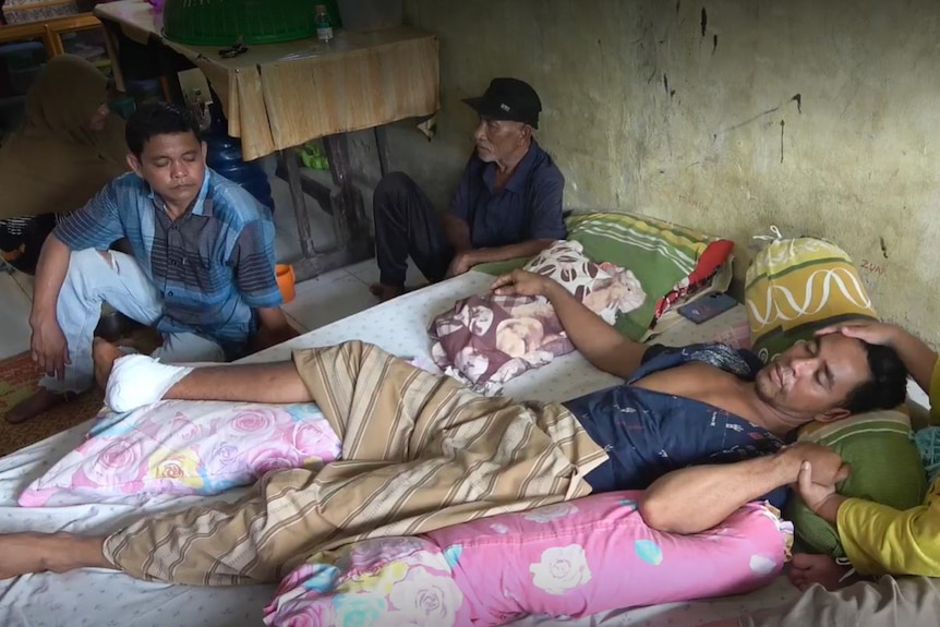 An Indonesian man lies on a hospital bed with an IV in his wrist and his bandaged foot resting on a pillow.