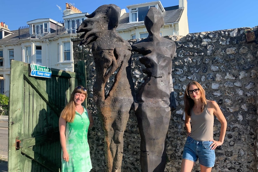 A modern stone statue of two women with two real women posing next to it