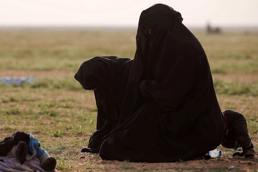 Two women in black niqabs kneel on the ground in prayer position.