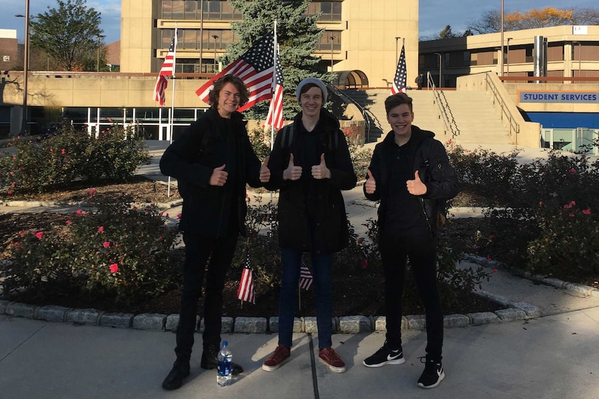 Three teenaged males, in winter clothing, smile and give the thumbs up outside a school building with American flags raised.