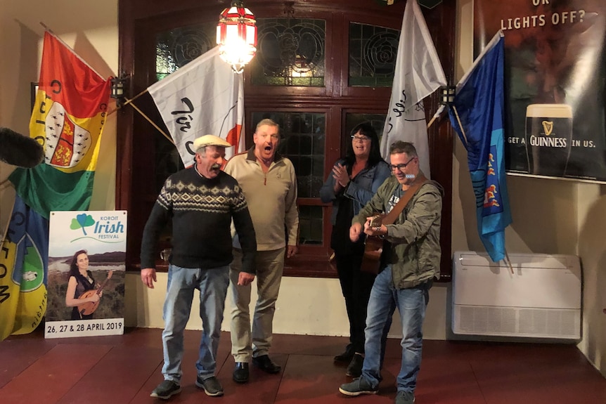 3 men and a woman singing and playing instruments in a large old fashioned entrance hallway of a pub. Irish flags in background.