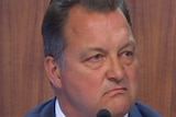 A man with brown hair scowls as he sits in a witness stand wearing a blue suit.