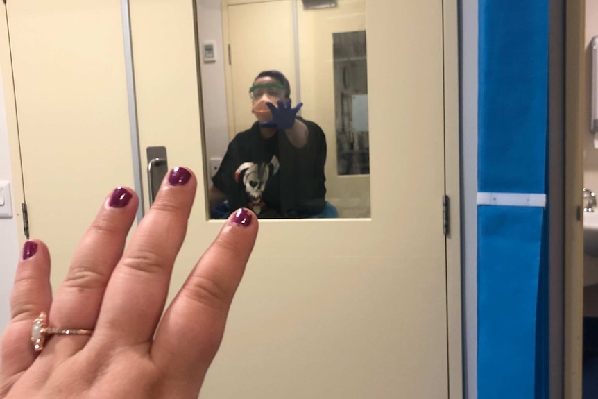 A woman in a mask looks through a glass door at a hospital and her partner's hand is visible reaching towards her