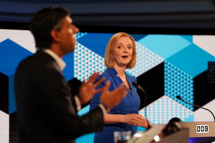 A man and a woman gesture as they stand behind podiums on a blue-lit stage.