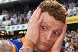 A male AFL player has his hands on both sides of his face as he stands looking at the ground.