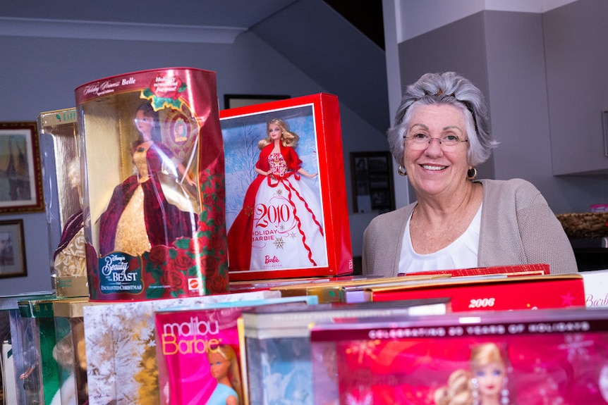 A woman smiles next to a range of Barbie dolls in boxes.