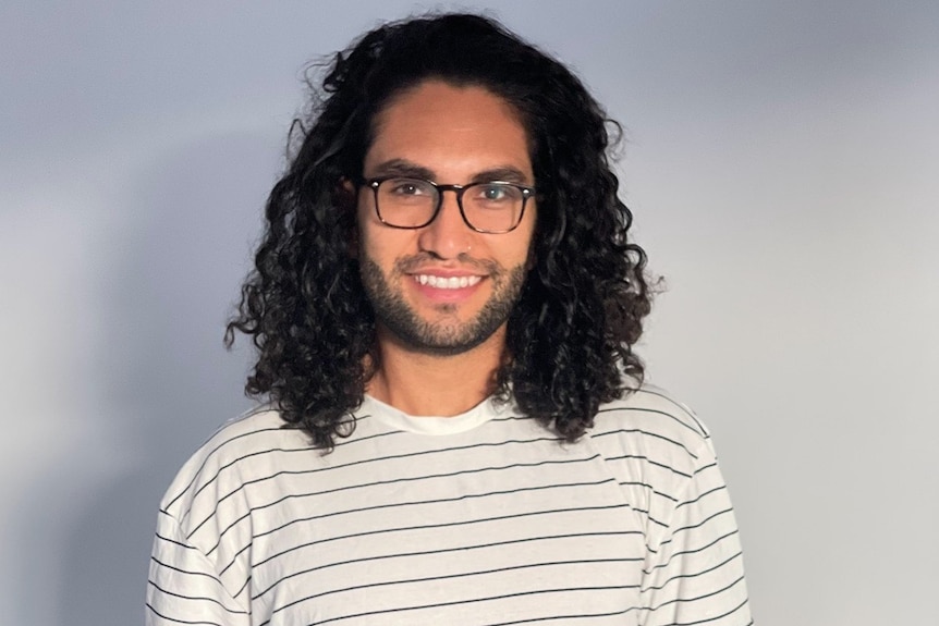 Man with shoulder-length black curly hair and glasses smiles at the camera. He wears a white and black striped shirt and jeans.