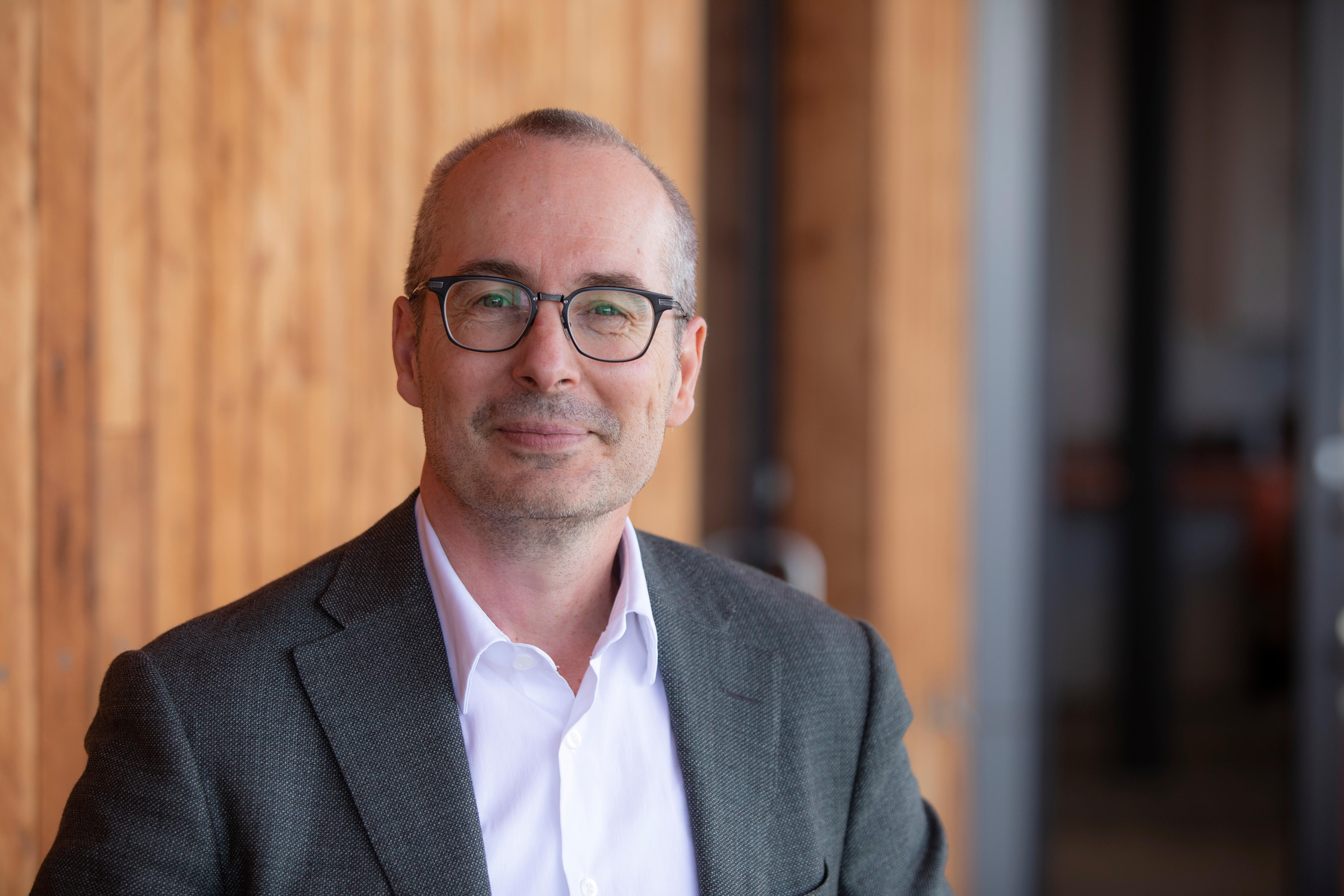 A bald man wearing a grey suit jacket and pink shirt with glasses smiling slightly