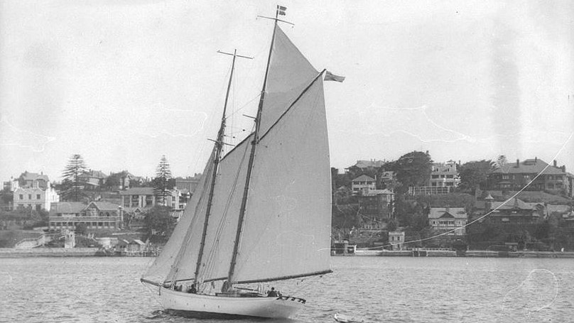 Black and white photo of a gaff rigged schooner under sail towing a dinghy, with a foreshore and houses behind it.