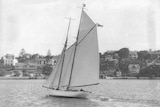 Black and white photo of a gaff rigged schooner under sail towing a dinghy, with a foreshore and houses behind it.