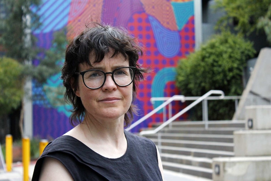 A middle aged woman with black hair stands in front of a colourful wall