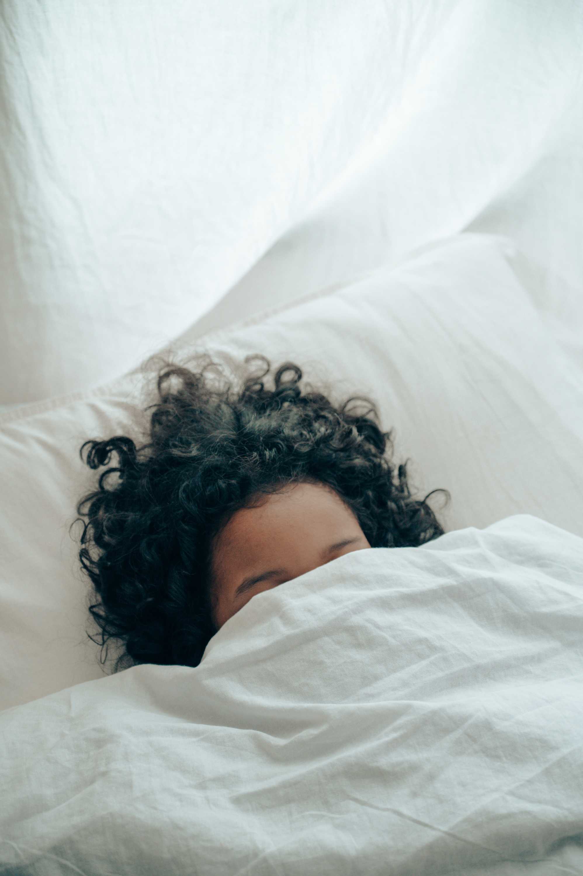 A woman with dark hair, hides her face behind a bed sheet.