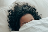 A woman with dark hair, hides her face behind a bed sheet.