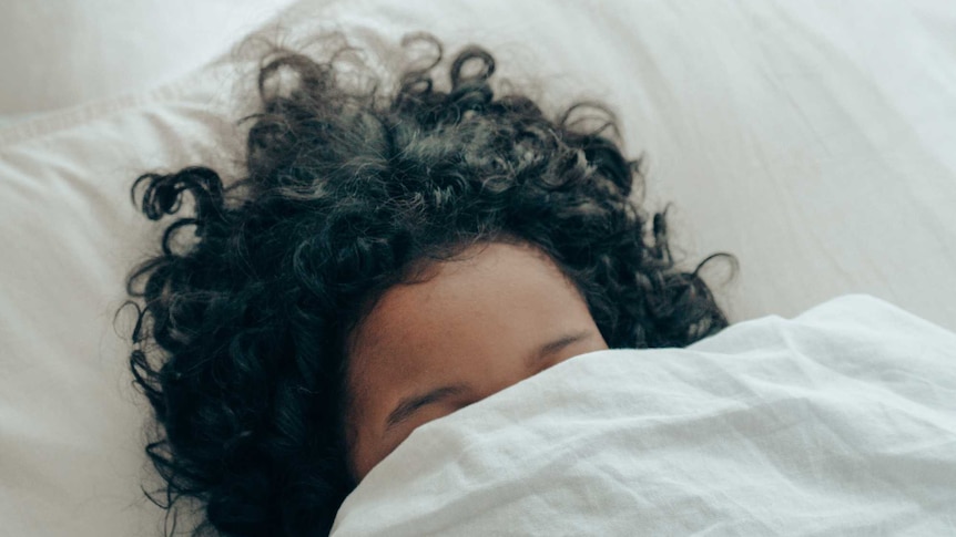 A woman with dark hair, hides her face behind a bed sheet.