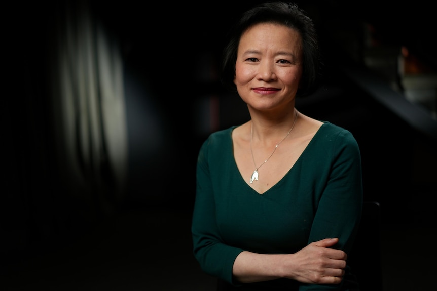 A middle-aged Australian woman of Chinese heritage smiles in front of a dark-curtained background.