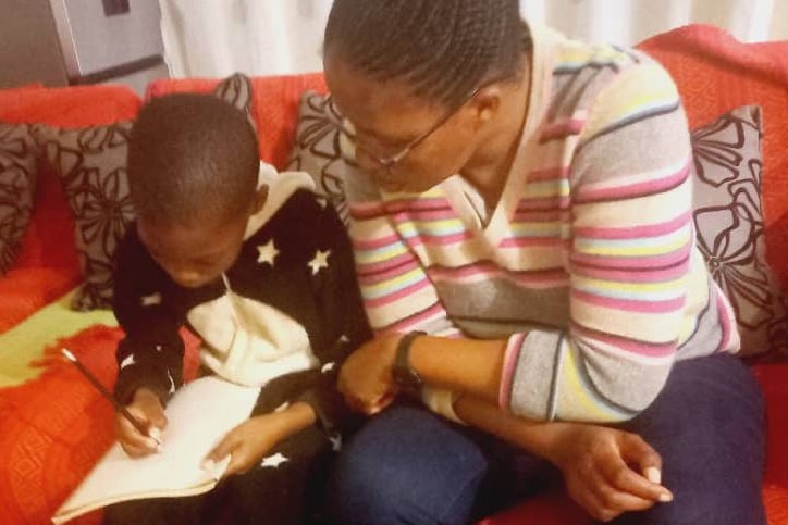 Child holds a pencil and writes on a piece of paper next to a woman wearing glasses.