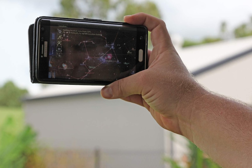 A man's hand holding a phone in the air. It's screen shows a map of a nearby galaxy.