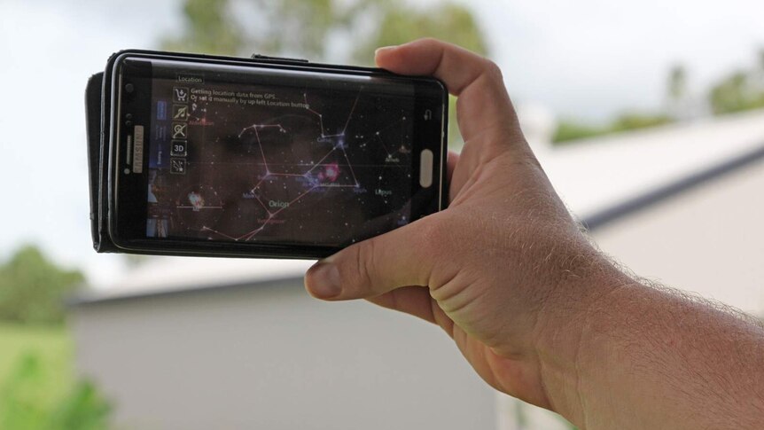 A man's hand holding a phone in the air. It's screen shows a map of a nearby galaxy.