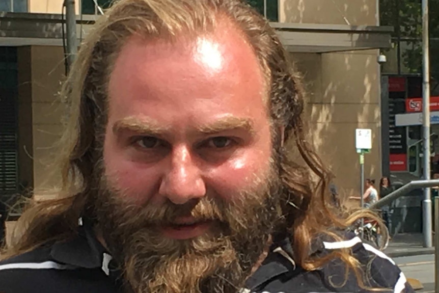 A bearded man with long hair looks at the camera.