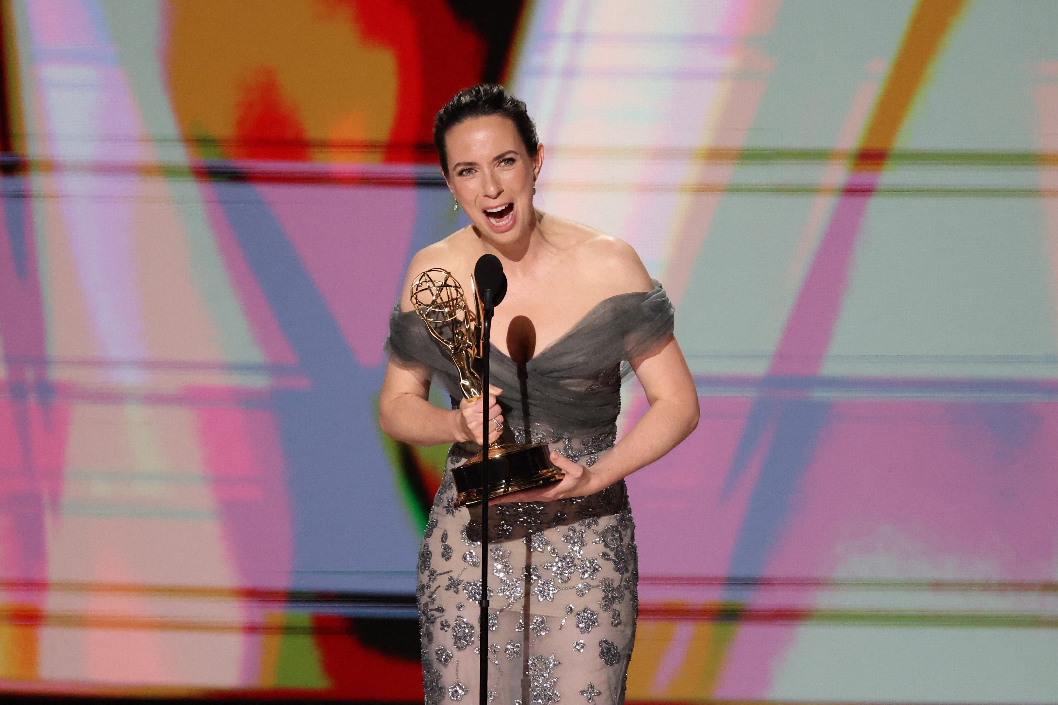 a woman in a gown holds emmy award at a mic stand