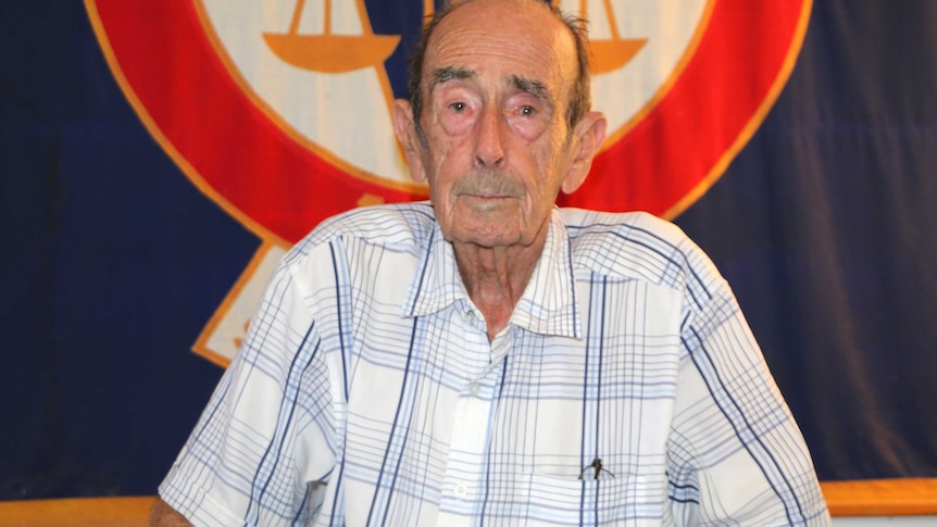 An old man poses for a photo sitting down in front of a blue and red flag.