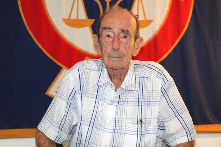 An old man poses for a photo sitting down in front of a blue and red flag.