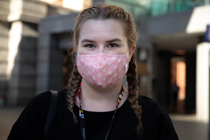 A woman with plaits wearing a pink face mask and a black top and a lanyard in Perth city.