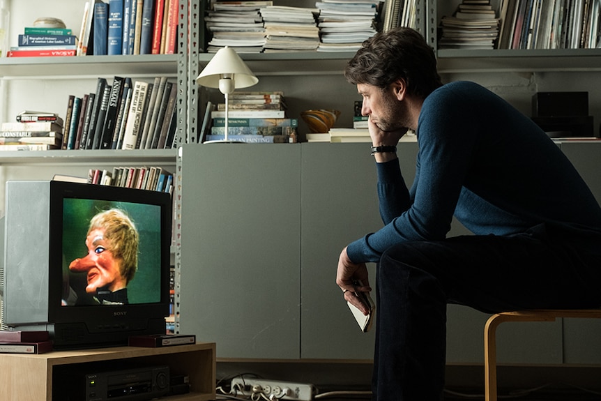 A man in navy pullover sits bent over on stool focused on small CRT television set next to a wall of grey metal bookshelves.